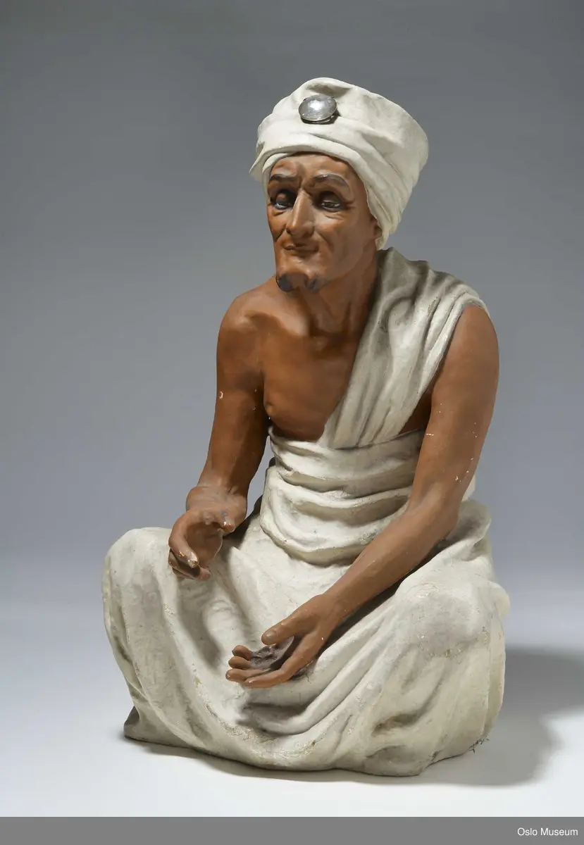Sittende helfigur, mann med "toga" og turban med imitert stor diamant. Utstrakt hånd, antagelig holdene tegneblyant.
Bilde nr 2: figuren er avbildet i lokalene til Schlytter og Wilhelmsen, kort tid før avvikling.