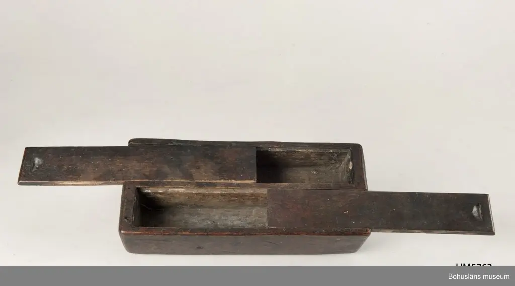 Rektangulär låda, uppdelad i två parallella fack med varsitt skjutlock upptill.
Målat med mörkt röd färg.
Inuti ett av facken finns fragment av fnöske.

Litt.; Gjaerder, Per, Esker og tiner, C. Huitfeldts förlag, Oslo, 1981, s. 48-50.

Ur handskrivna katalogen 1957-1958: 
Trälåda m. två skjutlock
a) L. 22,5 Br. 9,7, H. 5,3 cm, rödmålad; m. 2 fack, under m. ett skjutlock b) och c); deras L. 21,2 cm, Br 4 cm.

Lappkatalog: 84