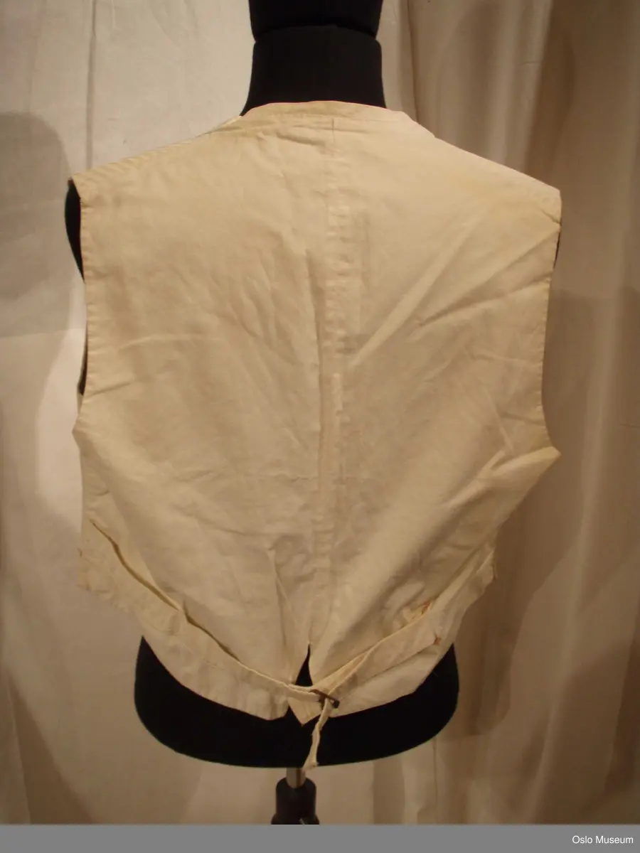 1: Vest i hvitt stoff med innvevde striper i brystparti. Knepping i form av 1 hvit knapp i perlemor, 3 mangler. Spenne bak.
2: Vest i hvitt stoff med innvevde ranker/blomster. Knepping foran med 3 hvite runde perlemor (?) - spenne bak