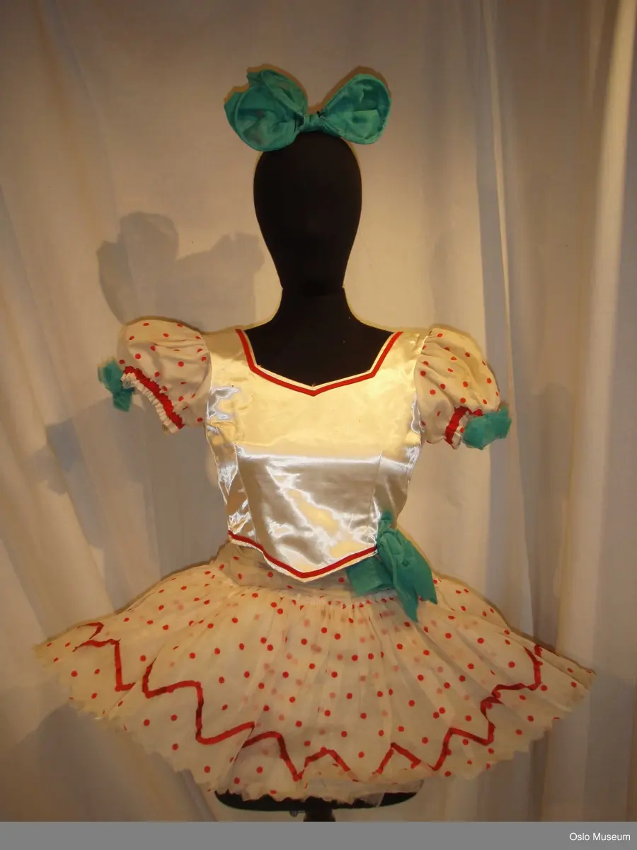 2 bluser i hvitt silkestoff med ermer av transparent, hvitt stoff med røde prikker. Røde kantebånd. Skjørtdel av hvitt tyllstoff med røde prikker og røde kantebånd nederst 2 stk.. 1 stor sløyfe med strikk i grønn tyll.