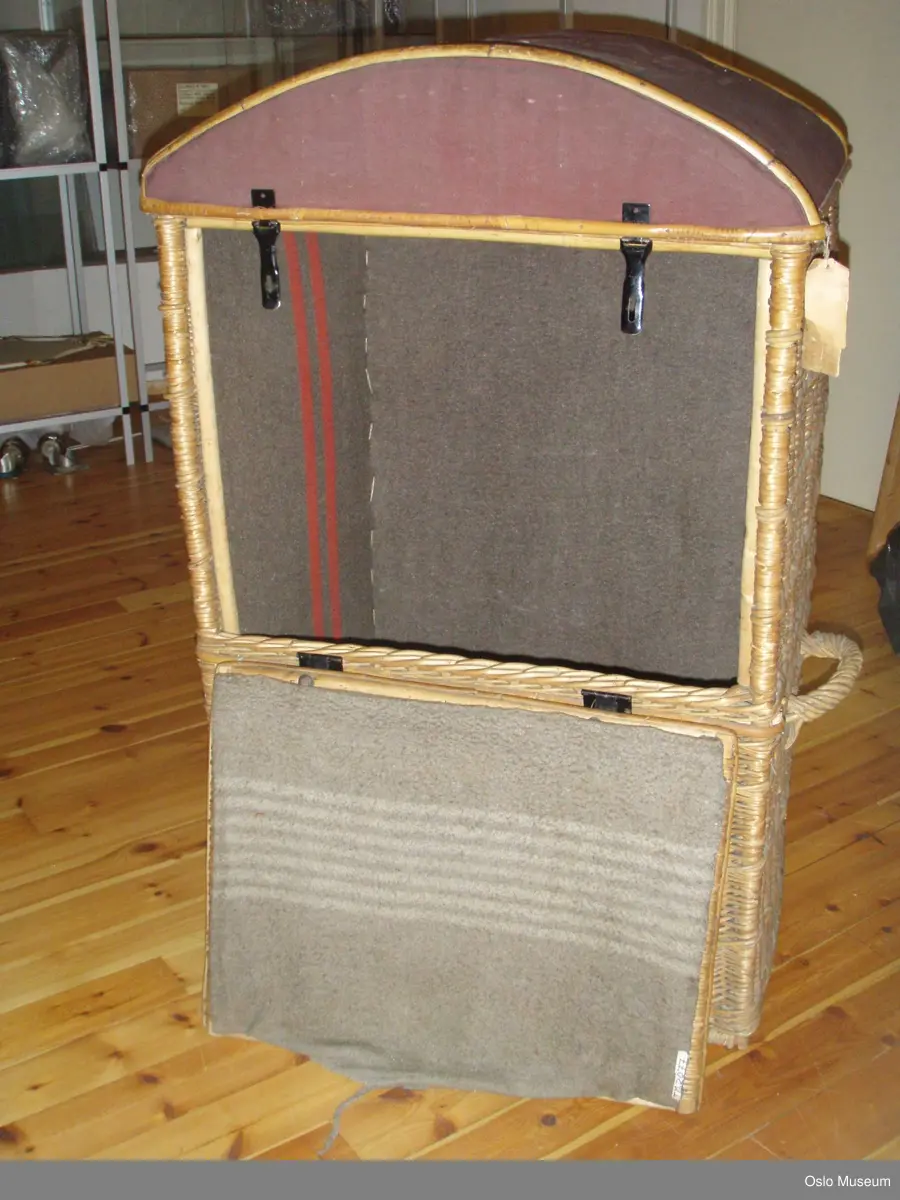 Reisegarderobe, amerikakoffert brukt til ballettutstyr i rotting med buet og lerret-trukket tak. 2 bærehanker. Åpning i front med beslag av metall. Innvendig trukket med ullstoff (ullteppe).