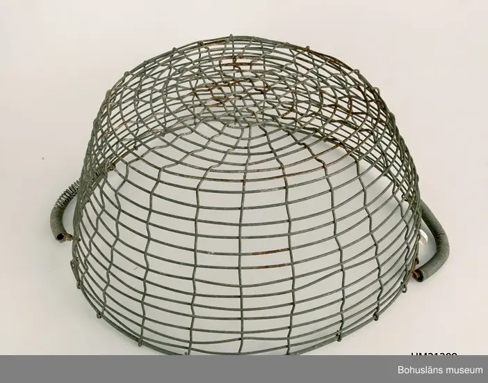 Rund korg vilken är svagt konande nedåt. Två ledade bärhandtag omvirade med ståltråd.
Korroderade.
Mått på den andra korgen: L 53,5, B 55, H 22 cm