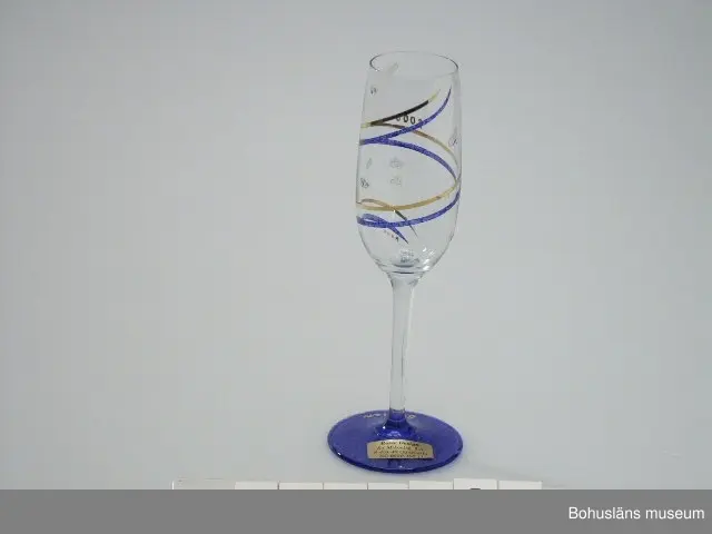 *Diam. avser kuppans bredaste del.

Smalt glas av klart glas på hög fot. Fotring av blåmarmorerat glas, kuppan dekorerad med två parallella linjer i guld och blått som löper runt glaset. Dekorer av strödda klisterstänk/klickar. Klistermärke på kuppan.

"Glas år 2000
Champagneglas använt av undertecknad på nyårsfest 1999/2000 hos bagarmästare Gösta Dahlskog och hans hustru Ulla, Bangatan 8 i Uddevalla, innehavare av Dahlskogs Bageri, Wienerkonditoriet och Princens konditori, alla i Uddevalla. 
Värdpar samt sju gäster. Varje glas hade försetts med respektive  gästs namn och "2000", liten reklamlapp från säljande affär, Rosie Design, Sankt Mikaelsgatan 2, Uddevalla. 
Detta glas användes under aftonen för dels vitt vin dels champagne (välkomst- och 12-slagstid). Till middag serverades färsk hummer, marulk som varmrätt och hjortronparfait till efterrätt. Efter 12-slaget avhörde värdpar och gäster ett stort antal större och mindre fyrverkerier.
Stellan Granat"

Materialet ingår i den Millennieinsamling som Bohusläns museum gjorde inför millennieskiftet 2000 på initiativ från SAMDOK och Nordiska museet. Insamlingsgruppen bevakade, samlade in respektive köpte under perioden närmast före och efter nyåret olika trycksaker, bl a Bibel 2000 i nyöversättning, dagstidningar från den 31 december 1999, postorderkataloger, samhällsinformation som fanns att köpa eller spreds till allmänheten av myndigheter. Flera av katalogerna avser att visa klädmode, leksaksutbud, matvanor etc. vid tiden för millennieskiftet.
Insamlingsgruppen gjorde personliga urval i butikerna i Uddevalla av det överflödande utbud av utrustning för firandet som särskilt tagits fram samt samlade in minnesföremål från det privata millenniefirandet. 

Som en kuriositet och hälsning till framtiden skrev Insamlingsgruppen också ett brev till kommande museikollegor på Bohusläns museum. 
Brevet är adresserat:
"TILL FRAMTIDEN
BOHUSLÄNS MUSEUM
BOX 803
451 19 UDDEVALLA"
frankerat och avsänt den 31 december 1999 och ankomststämplat av museets registrator  den 3 januari 2000. 
Brevet förvaras förseglat i sekretessskåpet i ämbetsarkivet och får ej öppnas förrän den 1 januari 2100.