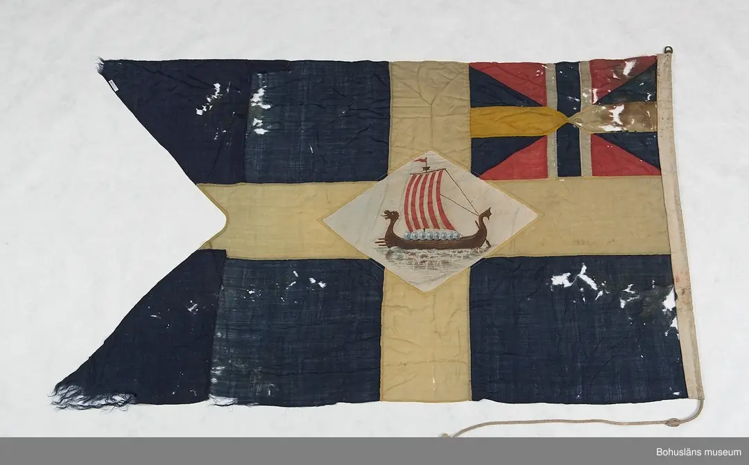 Segelsällskapet Vikens flagga, SS Viken. 
Svensk tvåtungad  flagga med parti "Silllsallaten", svensknorska unionsflaggan i övre hörnet samt infällt handmålat rombiskt emblem placerat mitt i det gula korset. På emblemet är målat ett vikingaskepp. 
Märkt "KASEN" med svart märkbläck. Hand- och maskinsömnad.
Skador. Eventuellt har flaggans form ändrats.