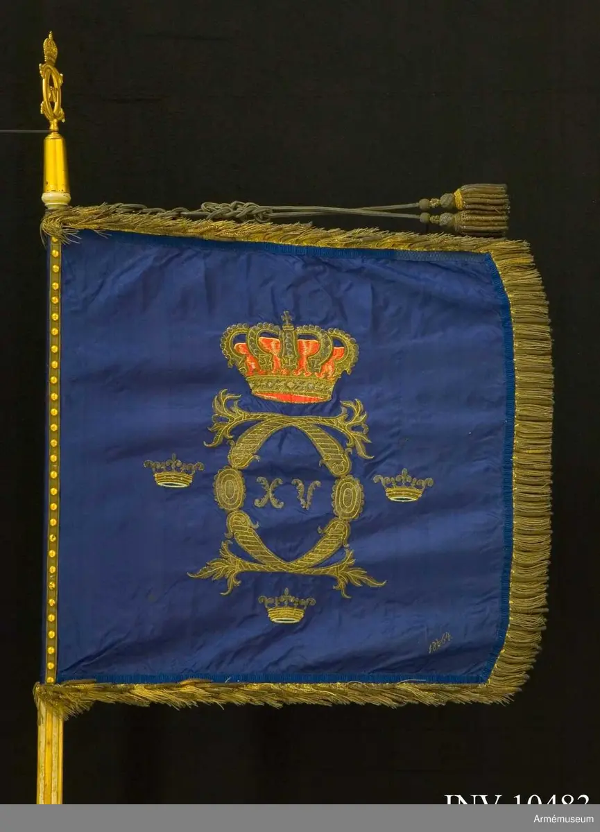 Grupp B I:2.

Duk av mörkblå (indigoblå) 4-skaftat fansiden med broderade emblem: CXVC. Rikt profilerade C under kunglig sluten krona med rött inner- och ytterfoder och silverstenar. Tre kronor med blått innerfoder i två nyanser. Alla broderier i guld. Kronans  foder rött fansiden med schattersöm. Frans av grövre och finare guldkantiljer. Signerad 18 3/5 64. Fastspikad med  guldbeläggningssnöre (majorssnöre). Kupiga tännlikor.
Stång: Rött med guld ovan duken, ränna för kravatt, doppsko  saknas. 660 mm, 1620 mm, 1780 mm, 2670 mm. Spets med Karl XV:s krönta monogram, l 152 mm. Holk 80 mm. Ränna 120 mm. Kravatt av gespinsterarbete, tofsar som fransen. Enkla knutar. Längd 1270 mm.