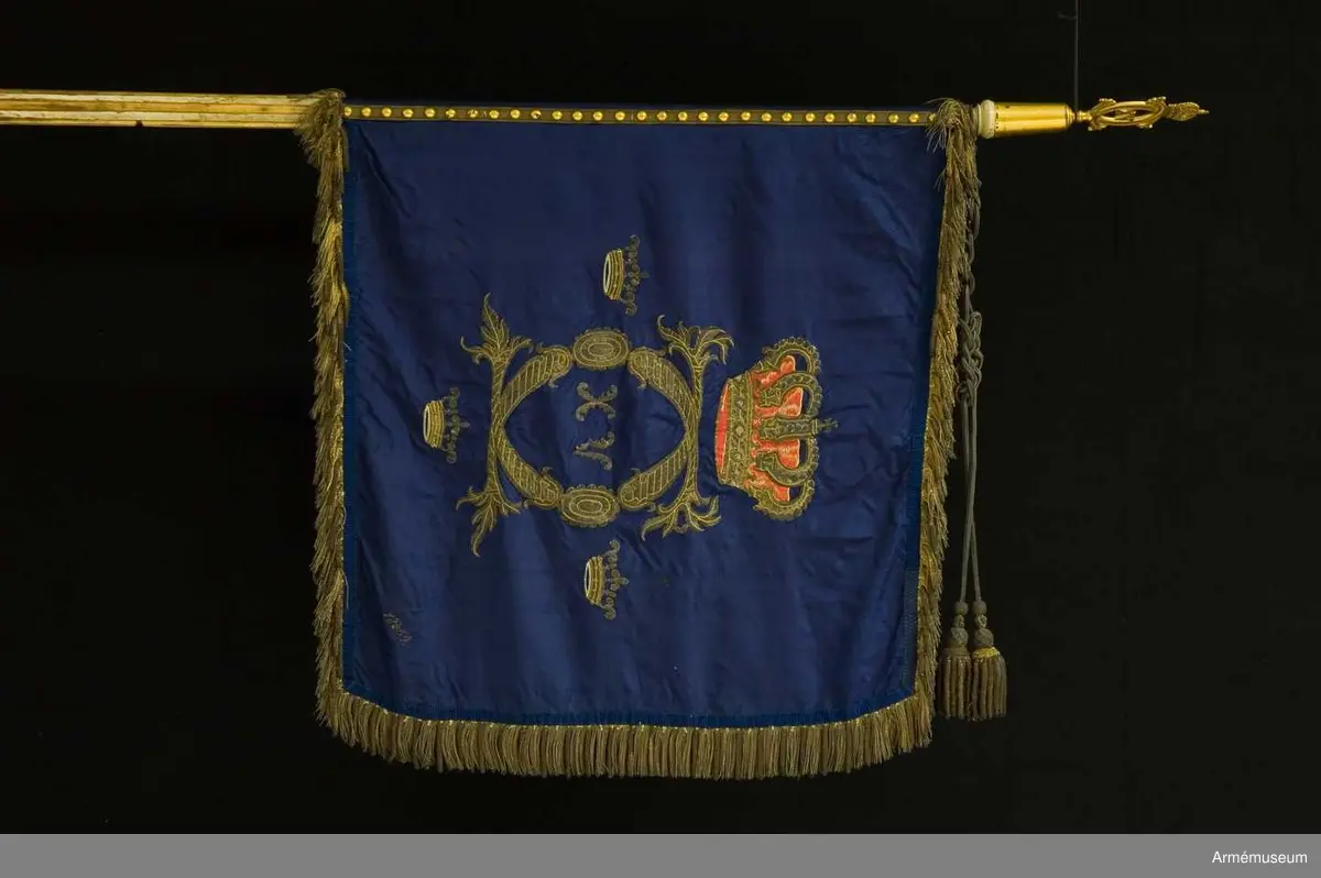 Grupp B I:2.

Duk av mörkblå (indigoblå) 4-skaftat fansiden med broderade emblem: CXVC. Rikt profilerade C under kunglig sluten krona med rött inner- och ytterfoder och silverstenar. Tre kronor med blått innerfoder i två nyanser. Alla broderier i guld. Kronans  foder rött fansiden med schattersöm. Frans av grövre och finare guldkantiljer. Signerad 18 3/5 64. Fastspikad med  guldbeläggningssnöre (majorssnöre). Kupiga tännlikor.
Stång: Rött med guld ovan duken, ränna för kravatt, doppsko  saknas. 660 mm, 1620 mm, 1780 mm, 2670 mm. Spets med Karl XV:s krönta monogram, l 152 mm. Holk 80 mm. Ränna 120 mm. Kravatt av gespinsterarbete, tofsar som fransen. Enkla knutar. Längd 1270 mm.