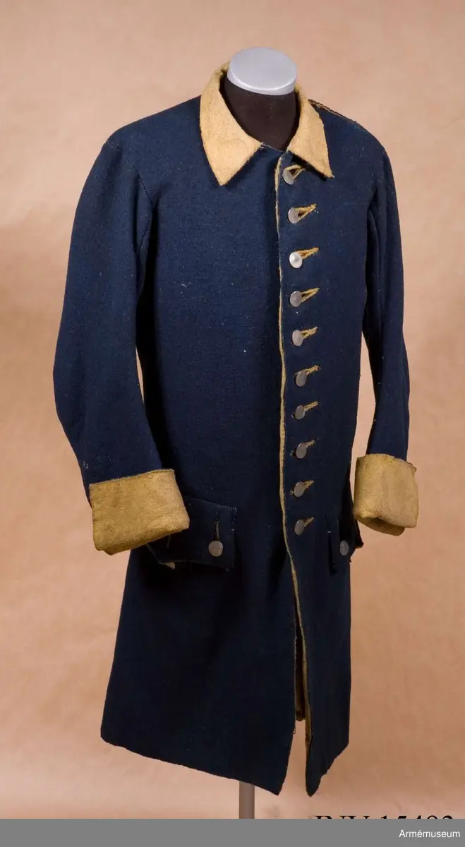 Rock av blått kläde med gul krage och ärmuppslag. Fodrad med gul boj i livet och med linne i ärmarna. Försedd med tennknappar.