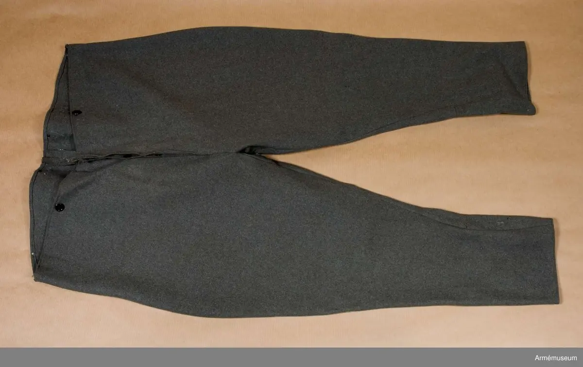 Av fältgrått tyg. De har sprund med tre knappar. För hängslen finns fyra knappar, många saknas.
