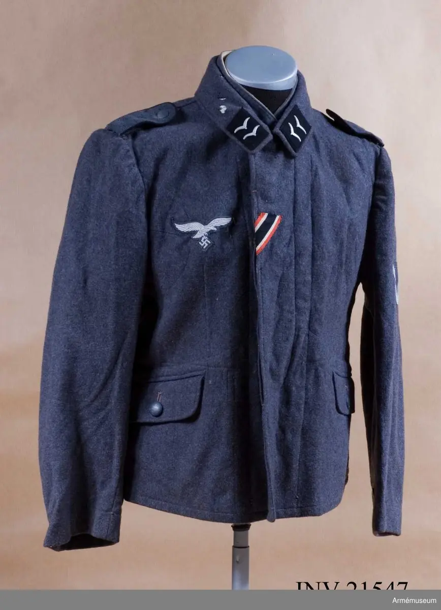 Av fältgrå kläde. Med knäppjulp. Axelkklaffar av samma grå kläde med svart passpoil. Foder av grått bomullstyg med två innerfickor. Knappar av grå metall. Krage, öppen, nedviken med klaffar av svart kläde på kragens ända.