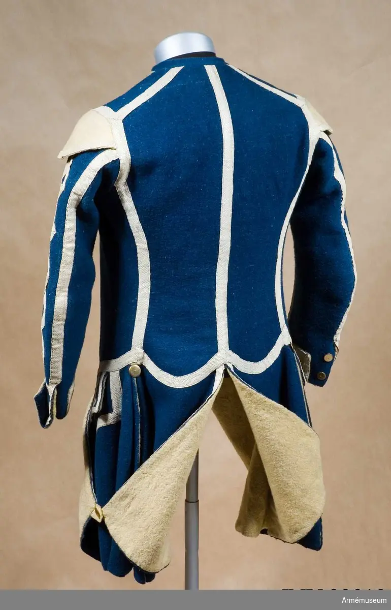 Rock med prydnader för spelet; mellanblå med gul rabatt (bröstrevärer), svalbon (axeldekoration) och ärmuppslag. Vit redgarnsdekor bruklig för spel. Rocken har sammanlagt 23 knappar. Knapparna i framkanten fastträdda med vitt band från avigsidan. En knapp i vardera skört (en ny). Två knappar på ficklocken (höger sida fattas en). Rocken är fodrad med gult ylletyg. Rocken ihopfästes med hyskar och hakar.