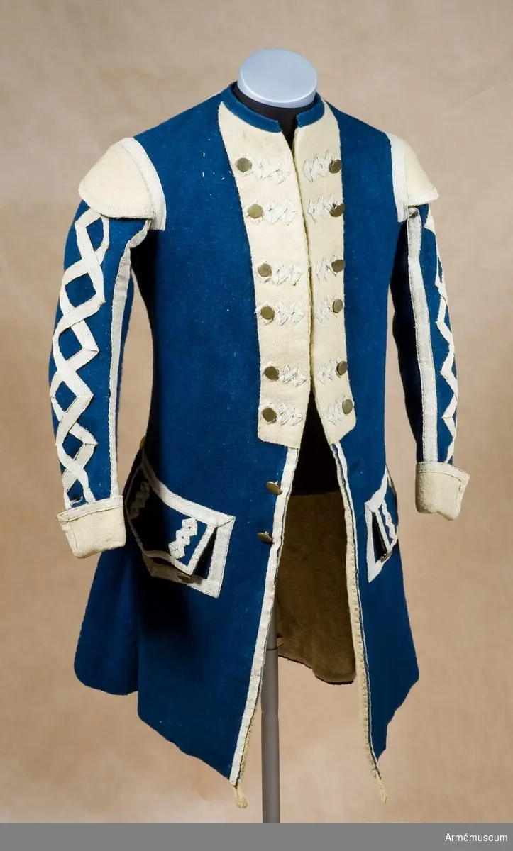 Rock med prydnader för spelet; mellanblå med gul rabatt (bröstrevärer), svalbon (axeldekoration) och ärmuppslag. Vit redgarnsdekor bruklig för spel. Rocken har sammanlagt 23 knappar. Knapparna i framkanten fastträdda med vitt band från avigsidan. En knapp i vardera skört (en ny). Två knappar på ficklocken (höger sida fattas en). Rocken är fodrad med gult ylletyg. Rocken ihopfästes med hyskar och hakar.