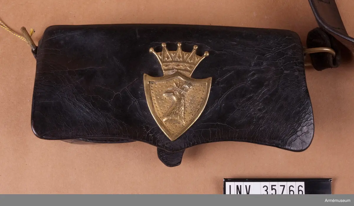 Kartuschväska av svart läder med mässingsbeslag i form av Skånes vapen. Kartuschen har en extra ficka för knallhattar och ett fack i botten som stängs med två remmar. De tillhörande spännena saknas. I kartuschen finns en träkloss med borrade hål med ca 18 mm diameter.