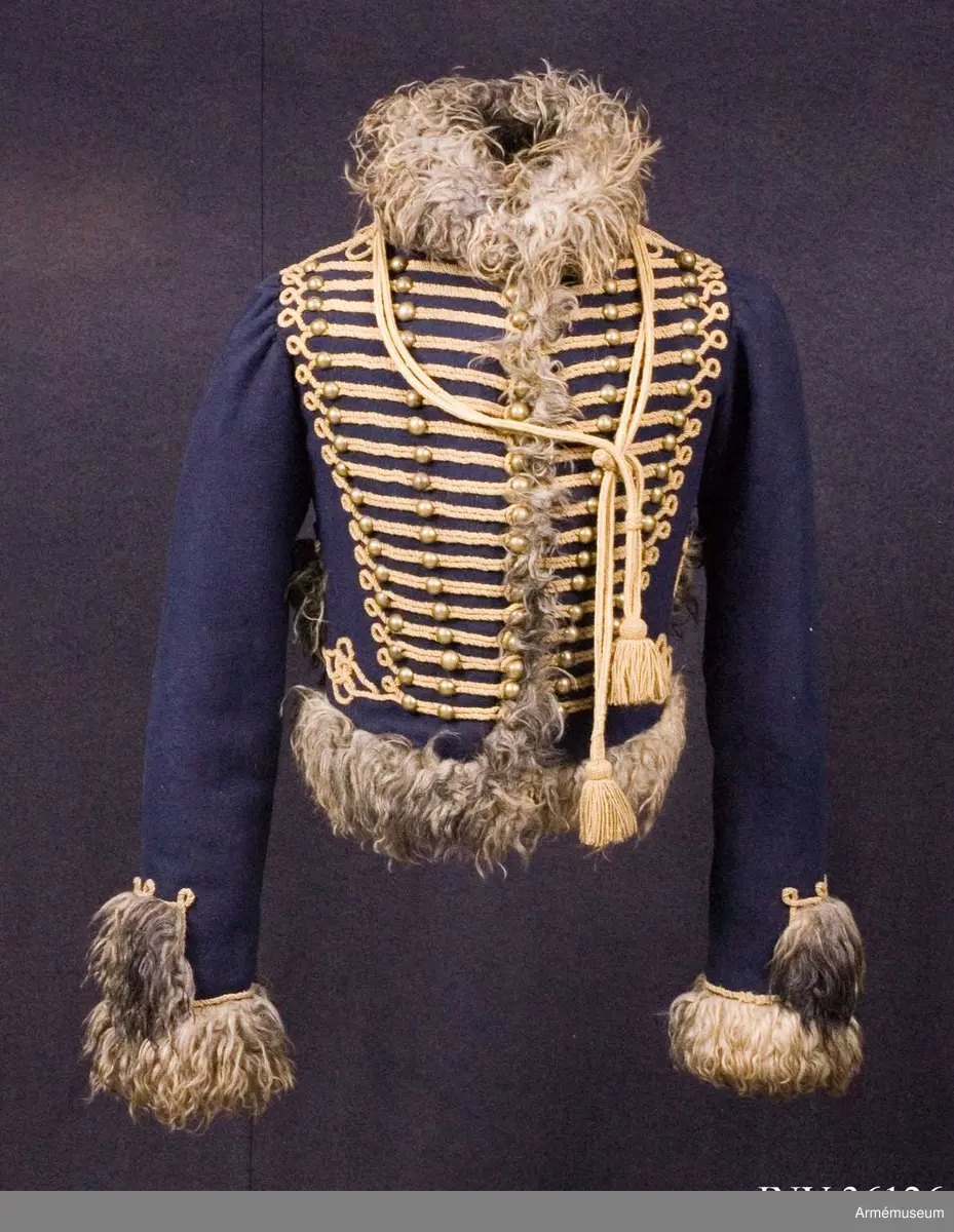 Blå päls med krage och ärmuppslag av gråbrunt fårskinn, med snören av gult ullgarn.

Pälsen är sydd av mörkblått kläde och är helfodrad med vitt, numera gulnat, korthårigt fårskinn. Ärmar, krage, framkanter och nederkant har bräm av gråbrunt fårskinn med lång lugg.
Plagget är konstruerat med fram- och sidstycken i ett samt helt bakstycke med litet skört i sidsömmarna. Ärmarna är ensömsärmar eller S-ärmar.
Tvärs över bröstet löper 18 rader snörmakerier gjorda av gul löst spunnen tvåtrådig ullsnodd. 5 knapprader, varav de två yttersta på varje sida har kullriga knappar medan knappraden i mitten som är avsedd att knäppas har klotknappar. Samtliga knappar är gjorda i en mässingslegering.
Ärmarna är kantade med päls. Pälsbrämet går upp i en "ruta" på framärmen, kantad med snodd. På baksidan av plagget nertill under ärmarna sitter två separata "rutor" av päls kantad med snodd. Sidsömmarna bak är markerade med snoddar som också pryder skörtets veck.
På baksidan av plagget nedanför kragen är det fäst ett flätat hängsle vars ändar hänger ner från varje axel och fram till plaggets främre nederkant. Varje del av hängslet är avslutat i en tofs. Vänster hängsle har ett spolformat knäppe klädd med snörmakeriarbete, medan det högra har en knut. Dessa är avsedda att hålla ihop plagget när det hänger öppet över vänster axel.
På vänster axel sitter en axelträns av dubbel snodd som sätts fast med en mässingsknapp.