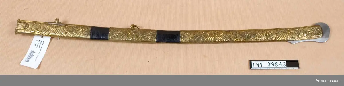 Grupp D II
Baljan har beslag, munbleck, mittbeslag och släpsko, av ursprungligen förgylld mässing. Alla tre beslagen är prydda med ciselerad, delvis graverad ornamentik. Släpskon har upptill urtagningar för styrskenornas nedre delar och baktill en med ränder prydd mässingsögla för en koppelrinvg. Öglan för den andra koppelringen sitter på mittbeslaget. Släpskon är längst ned på änden och på en bit av framkanten skodd med stål. Baljan påminner mycket om motsvarande del till sablar.