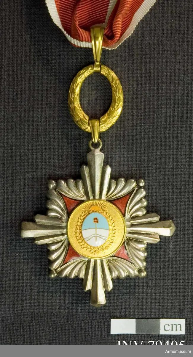 Kommendörstecken av argentinsk förtjänsteorden. På framsidan en man hållande argentinas fana i blått och vitt med texten "AL MERITO". På baksidan vapenskölden för Argentinas president.

Med band i rött med vita kanter.