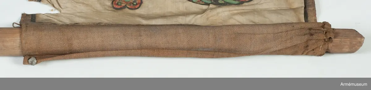 Duk av vit sidentaft och strumpa av rött linne. Motivet målat i guld- och silver samt rött och grönt bestående av en krönt örn, måne, sol och stjärnor, På duken en pappersetikett som anger att den tagits i slaget vid Saladen 1703.