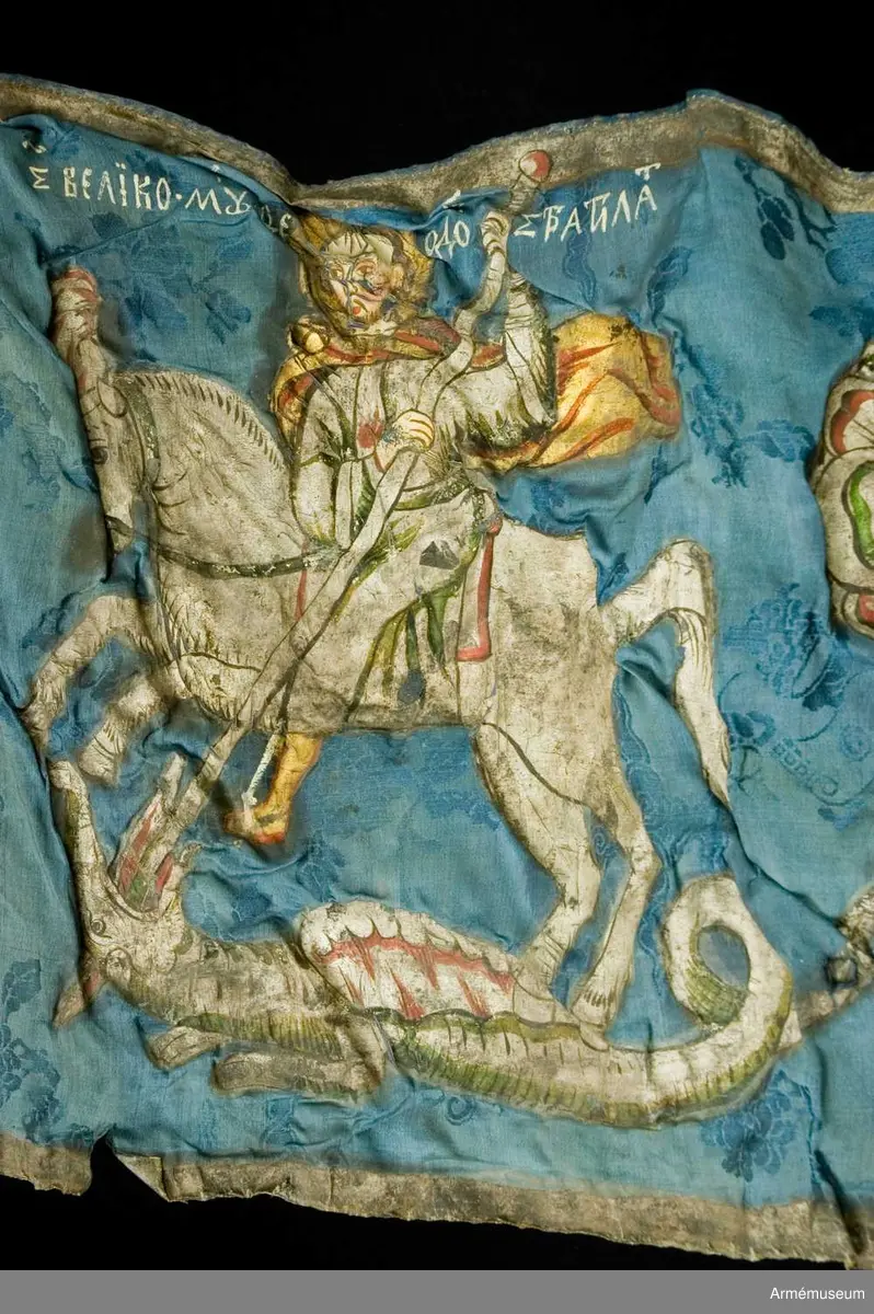 Entungad lansfana. Duk av blå kinesisk sidendamast med målad dekor. I 1703 års inventarium anges motivet vara St Göran men enligt den ryska inskriptionen är det Feodor Stratilat . Rosor och stjärnor av silver. Duken kantad med en silverbård. Strumpa av rött linne.