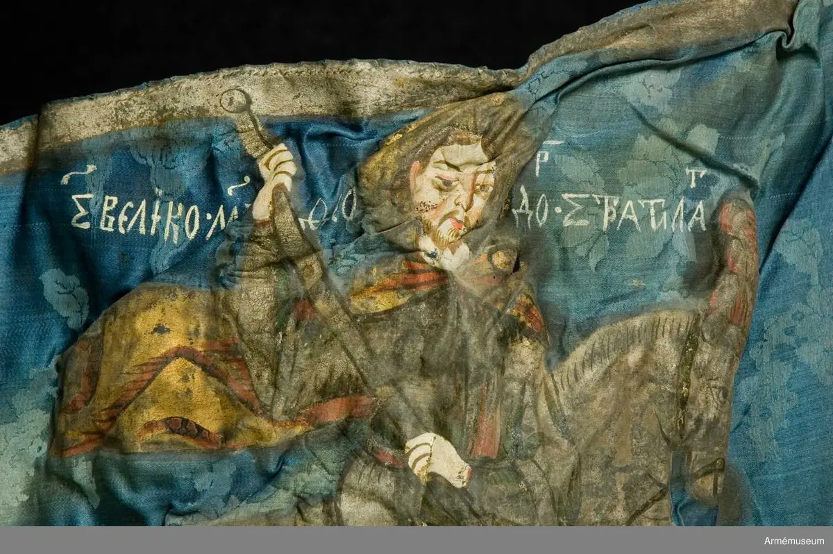 Entungad lansfana. Duk av blå kinesisk sidendamast med målad dekor. I 1703 års inventarium anges motivet vara St Göran men enligt den ryska inskriptionen är det Feodor Stratilat . Rosor och stjärnor av silver. Duken kantad med en silverbård. Strumpa av rött linne.