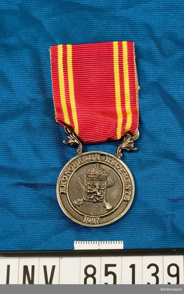 Medalj, minnes-, Kronobergs regemente (I 11). Medaljen är rund. Vapenskölden lagd över två korslgda musköter och krönt med en kunglig krona. I skölden ett lejon som bär ett armbost. På frånsidan en eklövskrans. Rött band med två smala gula band på vardera sidan.