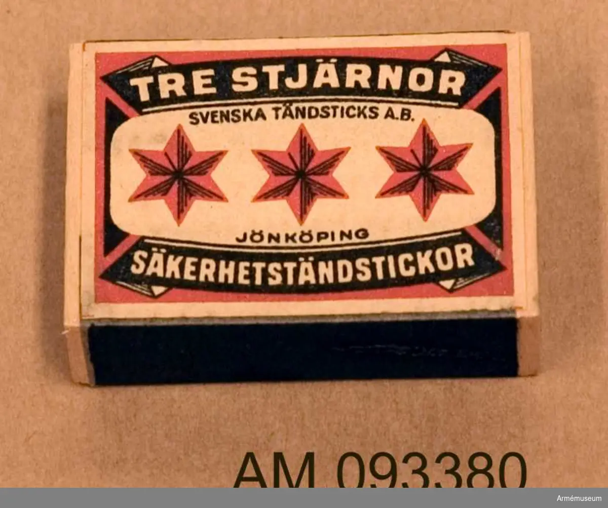 Tändsticksask med tändstickor. På ena sidan motivet Tre stjärnor och på andra sidan ett motiv som illustrerar hur en spionhand sträcker sig över en industribyggnad nattetid. 