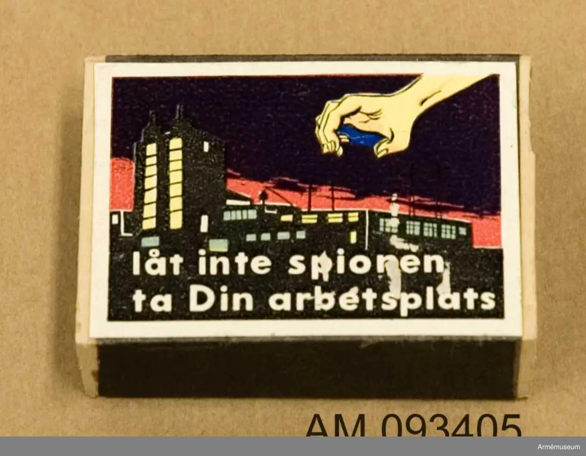 Tändsticksask med tändstickor. På ena sidan motivet Solstickan och på andra sidan ett klistermärke med motivet en spionhand som sträcker sig över en industribyggnad nattetid.
