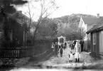 Ett antal barn och vuxna poserar längs en stensatt byväg. Mannen med kubben är folkskoleläraren Karl Bratt. Närmast i bild står hans brorsbarn, Maria och hennes lillasyster Sara.
I bakgrunden syns en stenmur med en portal. Där bakom syns ett högt berg.