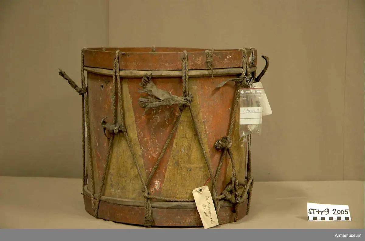 Cylindertrumma av trä med bemålad lim och krita grunderad duk.
Ytan är krackelerad med ett visst bortfall, dock relativt stabil. Tre hål i varierande storlek.

Membran: övre skinnet är smutsigt och repigt med tendenser till sprickbildning. Det finns en centimeterstor lagning. Det undre, har en tvärsgående spricka som tudelar skinnet. Den större delen rullar in mot mitten. Det finns fläckar av ngn. lättlöslig, gråaktig lösning. 

Veckelgjordar: har lindade fogar, den övre med tunt rep/lina, 
Den undre med rep och smalt läderband. Tre tecken inskurna i övre veckelgjordens utsida, som liknar: 1:a,  gaffel med tre klor, 2:a, endast de två yttre klorna och skaft, 3:e tecknet: endast de två yttre klorna, skaftet saknas. 

Spännlina: är fin med tvinnat, avsmalnande repslut. Det finns åtta spännstroppar varav två stycken är fransade.

Spännringar : Utsmyckning : röda kilar målade på gul botten. Mönstret ristat i grunden.

Märkning: fastklistrad Konung Karl den XII med ifyllnad: Litt B  No.4 
Hängandes pappers etikett  i påse No 1211,
Gustavianska koret No 9.