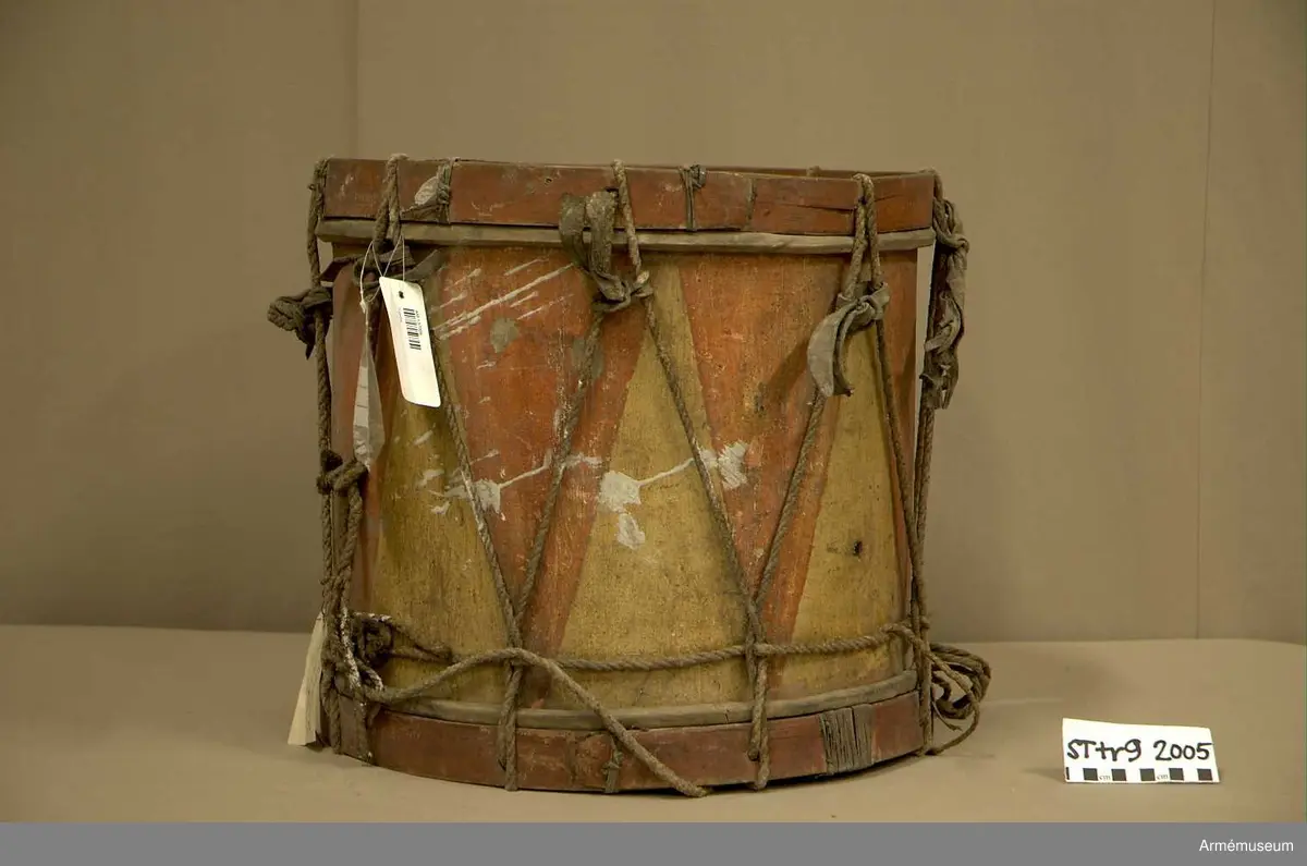 Cylindertrumma av trä med bemålad lim och krita grunderad duk.
Ytan är krackelerad med ett visst bortfall, dock relativt stabil. Tre hål i varierande storlek.

Membran: övre skinnet är smutsigt och repigt med tendenser till sprickbildning. Det finns en centimeterstor lagning. Det undre, har en tvärsgående spricka som tudelar skinnet. Den större delen rullar in mot mitten. Det finns fläckar av ngn. lättlöslig, gråaktig lösning. 

Veckelgjordar: har lindade fogar, den övre med tunt rep/lina, 
Den undre med rep och smalt läderband. Tre tecken inskurna i övre veckelgjordens utsida, som liknar: 1:a,  gaffel med tre klor, 2:a, endast de två yttre klorna och skaft, 3:e tecknet: endast de två yttre klorna, skaftet saknas. 

Spännlina: är fin med tvinnat, avsmalnande repslut. Det finns åtta spännstroppar varav två stycken är fransade.

Spännringar : Utsmyckning : röda kilar målade på gul botten. Mönstret ristat i grunden.

Märkning: fastklistrad Konung Karl den XII med ifyllnad: Litt B  No.4 
Hängandes pappers etikett  i påse No 1211,
Gustavianska koret No 9.