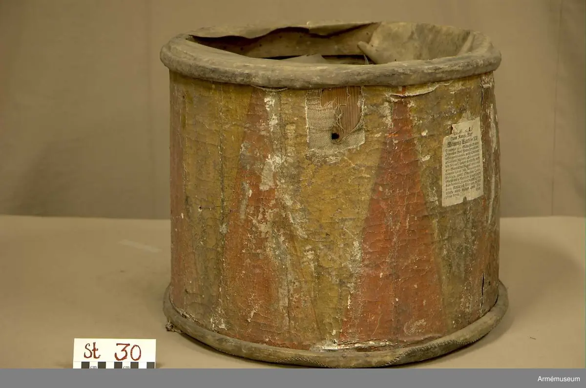 Cylindertrumma av: trä därpå spänd lim o krita grunderad duk som är bemålad. Hål i olika storlek, i jämn rad efter hela sidan. Svårt att säga stick eller insekts hål. Dessutom 7 st. hål i olika storlek, placerade runt om på cylindern. Till höger om den klistrade etiketten finns en stor ruta ca 8x7 cm. med ett totalt bortfall av kredering. För övrigt är ytan mkt. krackelerad och ostabil. 

Membran : övre skinnet är trasigt, undre skinnet är fogat efter spännringen och vågigt.

Veckelgjordar: saknas.

Spännlina: saknas. 

Spännringar : två stycken.

Utsmyckning : bemålad med röda kilar på gul botten. Ristat mönster. 

Märkning : klistrad färdigtryckt Konung Carl den XII etikett med ifyllnad
Litt. B. -No 25.