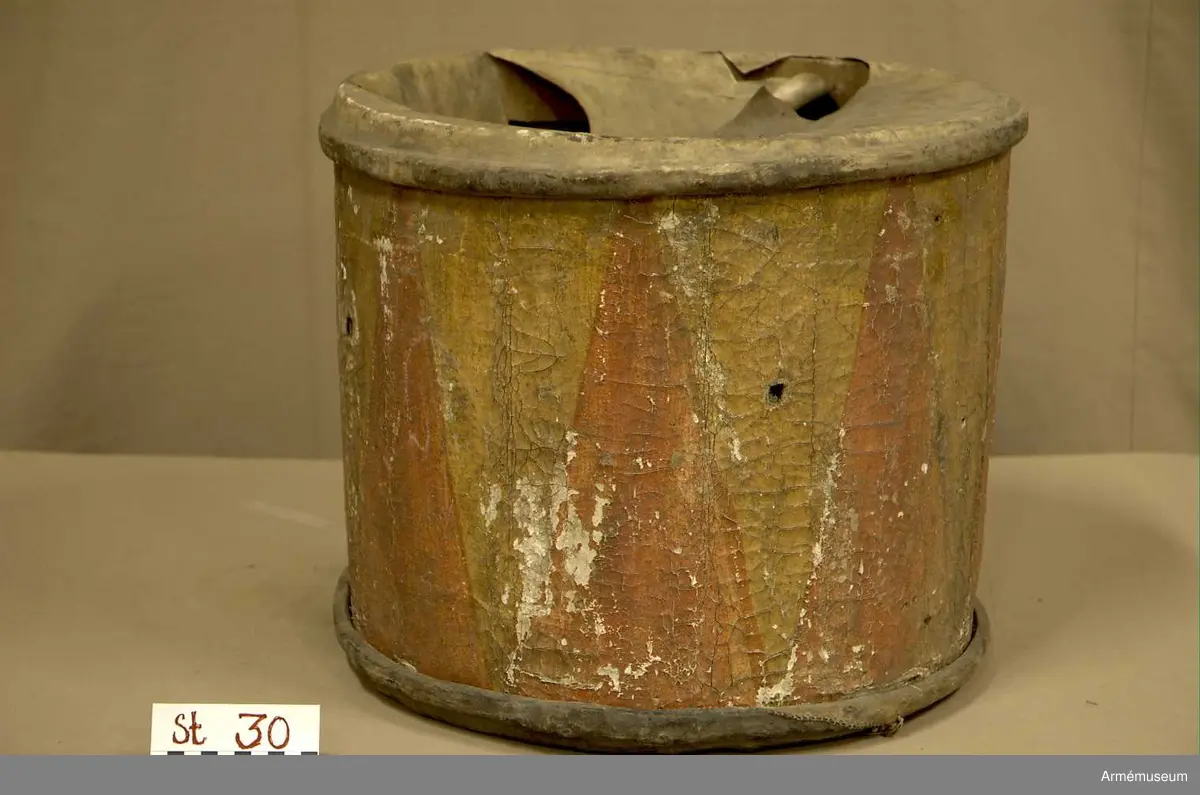 Cylindertrumma av: trä därpå spänd lim o krita grunderad duk som är bemålad. Hål i olika storlek, i jämn rad efter hela sidan. Svårt att säga stick eller insekts hål. Dessutom 7 st. hål i olika storlek, placerade runt om på cylindern. Till höger om den klistrade etiketten finns en stor ruta ca 8x7 cm. med ett totalt bortfall av kredering. För övrigt är ytan mkt. krackelerad och ostabil. 

Membran : övre skinnet är trasigt, undre skinnet är fogat efter spännringen och vågigt.

Veckelgjordar: saknas.

Spännlina: saknas. 

Spännringar : två stycken.

Utsmyckning : bemålad med röda kilar på gul botten. Ristat mönster. 

Märkning : klistrad färdigtryckt Konung Carl den XII etikett med ifyllnad
Litt. B. -No 25.