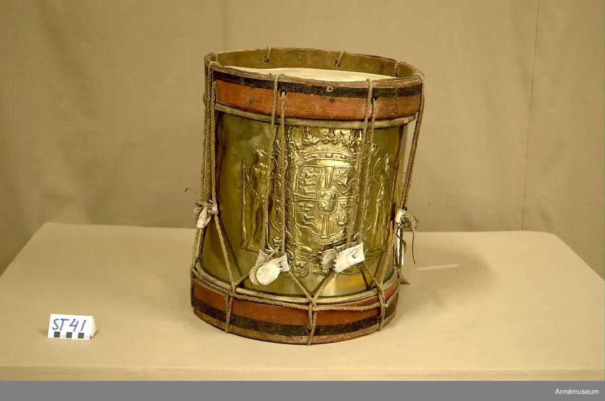 Cylindertrumma av : ljus metall, dekorerad. Metallen är bucklig och har ett mindre hål.

Membran : båda skinnen är trasiga. Ovanpå det övre skinnet ligger en färgad pappskiva. Det undre skinnet har en skada, ca 4 cm. med en flik som går inåt.

Veckelgjordar : träet skiktar sig i den övre veckelgjorden.
 
Spännlina : ser hel ut med 8 st. spännstroppar av vitfärgade tjocka  läderband ca 3 cm. breda. Tre av dessa har tandklippta avslut. 

Spännringar : finns.

Bärring : 

Utsmyckning : vapensköld med mansfigurer på vardera sidan.

Märkning : finns.