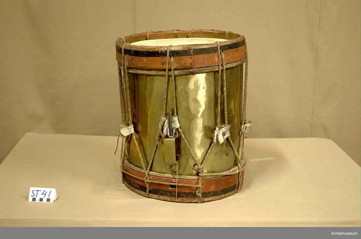 Cylindertrumma av : ljus metall, dekorerad. Metallen är bucklig och har ett mindre hål.

Membran : båda skinnen är trasiga. Ovanpå det övre skinnet ligger en färgad pappskiva. Det undre skinnet har en skada, ca 4 cm. med en flik som går inåt.

Veckelgjordar : träet skiktar sig i den övre veckelgjorden.
 
Spännlina : ser hel ut med 8 st. spännstroppar av vitfärgade tjocka  läderband ca 3 cm. breda. Tre av dessa har tandklippta avslut. 

Spännringar : finns.

Bärring : 

Utsmyckning : vapensköld med mansfigurer på vardera sidan.

Märkning : finns.