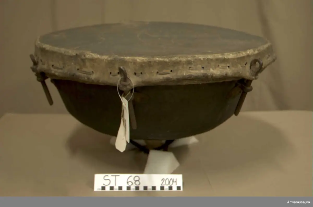 Kittel av koppar och skinn av kalv med fast spännring. Tredelad fot av järn. 7 skruvar och skruvfästen av järn. Bärrem av kalvskinn i två lager med spänne av järn.