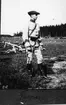 Soldat klädd i uniform m/1910 stående på stort fält, andra soldater ligger i bakgrunden. Fotot betitlat 