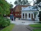 Järnhandlarhuset på Skansen fotograferad från en plats till vänster om Rosenträdgården. Fotografiet visar Järnhandlarhusets framsida med gatuplanet framför där några Vitkindade gäss, Skansens gäster och klädda pedagoger rör sig. I bakgrunden syns Boktryckarhusetett och längst till vänster skymtar Tottiska gården.