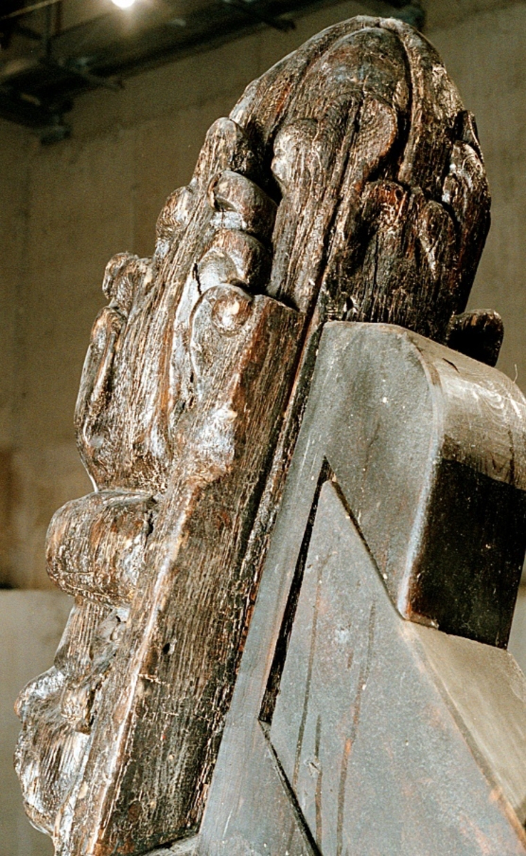 Skulptur i form av en hög, rikt snidad pelare eller stolpe, med kvadratisk genomskärning.

Tre av pelarens sidor är skulpterade medan den fjärde utgör anliggningsyta, försedd med ett antal olikformade urtag. De skulpterade sidorna är horisontellt indelade i tre eller fyra sektioner eller nivåer, avgränsade från varandra av profilerade och snedställda vulster. Nedtill avslutas skulpturen av en ornerad och på mitten insvängd formation.



Ovanför denna nedre avslutning är pelaren utformad som en reslig mansfigur, vänd mot vänster, sedd från betraktaren. Mansfiguren, som täcker tre av skulpturens fyra sidor, är iförd en romersk rustning med en bred fältbindel draperad från höger axel snett över bröstet. På huvudet med det lockiga och utslagna håret sitter en krona av öppen typ, med liljeliknande ornament. Figurens vänstra arm är böjd, medan den högra armen är sträckt utmed sidan. Handen till den högra armen saknas, men har hållit en rikt profilerad sköld, med mycket slitna och vaga konturer av en på bakbenen mot vänster gående grip med stora vingar. Skölden är snidad i ett med figuren, men med kraftiga underskärningar. Vid figurens fötter skymtar ett djur av något slag. Bakom mannens huvud tycks ett snidat textilstycke, kanske del till ett draperi, vara anbragt.



Ovanför den romerska krigaren eller riddaren har pelaren formen av en herm utan armar med långt, krusigt hår och mustaschprytt ansikte. Liksom mansfiguren täcker även hermen tre sidor av pelaren. Huvudet är vridet mot vänster. Överkroppen är bar så när som på ett flikigt plagg som täcker axlar och övre delen av bröstet. Från huvudet och runt midjan är ett tygstycke draperat. Hermens nederdel är formad till en stor maskaron med uppspärrad mun och utsträckt tunga. Maskaronen övergår upptill och utmed sidorna i yppiga bladformationer.



Pelaren avslutas upptill av ett knoppliknande ornament. Ornamentet är ornerat med akantusbladliknande bildningar samt fyra byglar som möts på toppen, där vissa spår antyder att det ursprungligen suttit ett klot eller dylikt, men som fallit bort. Eventuellt kan ornamentet utgöra en huvudbonad till hermen.

Skulpturen är kraftigt sliten.



Text in English: Sculpture in the shape of a richly carved column or pillar.

Three sides of the pillar are carved, while the fourth side comprises the attachment surface and has a number of variously-shaped indentations. The sculptured sides are divided horizontally into three or four sections separated by angled tori.



The lower section of the pillar is carved in the figure of a tall man. The figure is clad in Roman armour and has a field badge (cloak/plaid) draped across his chest at an angle from his right shoulder. On his head he wears an open crown. He has long, curly hair. The right arm is broken off, the left is at his side. The left hand is missing but once held a richly carved shield.

The shield is carved out of the same piece of wood as the figure, but deeply undercut. A carved figure is apparent on the shield. Although the shield is badly worn, it is possible to recognise one of the hind legs of a griffin with large wings. At the man's feet lies an animal of some kind.



Above the Roman warrior or knight, the pillar is carved in the shape of a herm, without arms and with long, curly hair and a moustache. Like the male figure, the herm also covers three sides of the pillar. The head is twisted to the left. The upper part of the body is bare except for a piece of cloth that covers the shoulders and upper chest. The lower part of the herm is carved as a large mask, its mouth open and tongue protruding. The sides and upper parts of the mask are carved as luxuriant leaves.



The upper part of the pillar has a ornament in shape of a knob. The ornament has a ring of acanthus leaves and four spherical loops run vertically from the acanthus leaves and meet at a point at the top. An object, for example a globe appears to have adorned the top of the ornament. This object is missing. The ornament may also be a part of the headgear of the herm.

The sculpture is severely worn.