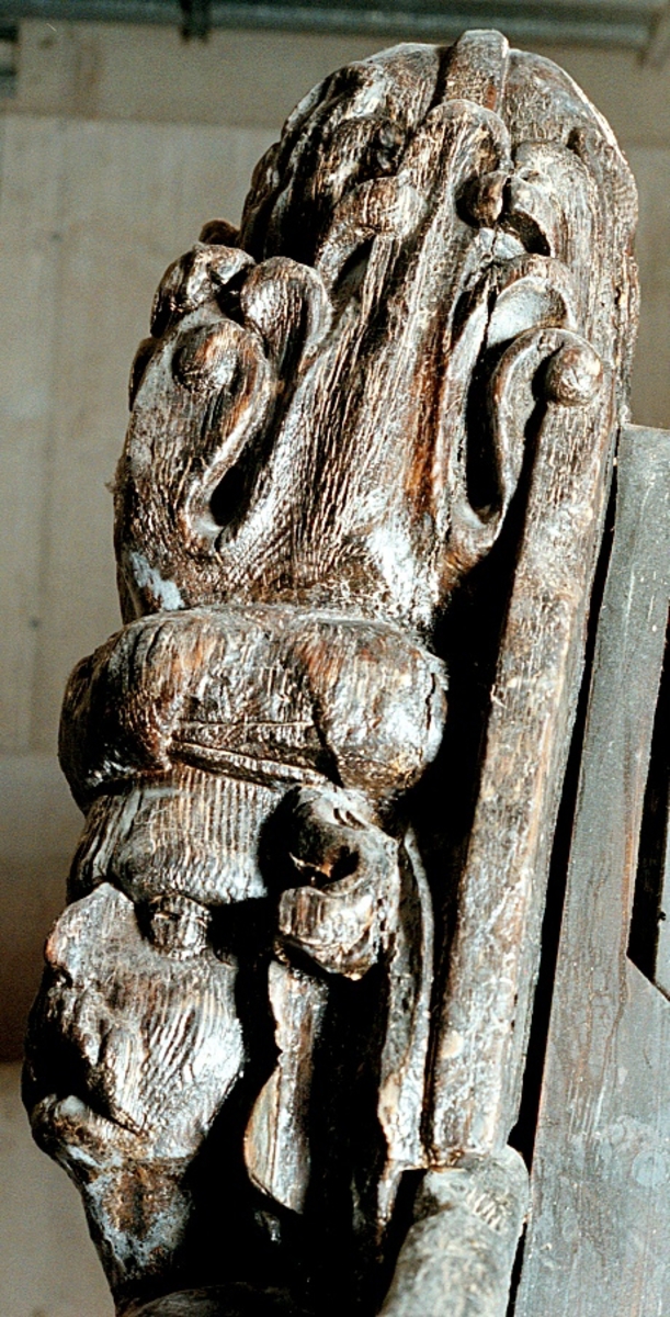 Skulptur i form av en hög, rikt snidad pelare eller stolpe, med kvadratisk genomskärning.

Tre av pelarens sidor är skulpterade medan den fjärde utgör anliggningsyta, försedd med ett antal olikformade urtag. De skulpterade sidorna är horisontellt indelade i tre eller fyra sektioner eller nivåer, avgränsade från varandra av profilerade och snedställda vulster. Nedtill avslutas skulpturen av en ornerad och på mitten insvängd formation.



Ovanför denna nedre avslutning är pelaren utformad som en reslig mansfigur, vänd mot vänster, sedd från betraktaren. Mansfiguren, som täcker tre av skulpturens fyra sidor, är iförd en romersk rustning med en bred fältbindel draperad från höger axel snett över bröstet. På huvudet med det lockiga och utslagna håret sitter en krona av öppen typ, med liljeliknande ornament. Figurens vänstra arm är böjd, medan den högra armen är sträckt utmed sidan. Handen till den högra armen saknas, men har hållit en rikt profilerad sköld, med mycket slitna och vaga konturer av en på bakbenen mot vänster gående grip med stora vingar. Skölden är snidad i ett med figuren, men med kraftiga underskärningar. Vid figurens fötter skymtar ett djur av något slag. Bakom mannens huvud tycks ett snidat textilstycke, kanske del till ett draperi, vara anbragt.



Ovanför den romerska krigaren eller riddaren har pelaren formen av en herm utan armar med långt, krusigt hår och mustaschprytt ansikte. Liksom mansfiguren täcker även hermen tre sidor av pelaren. Huvudet är vridet mot vänster. Överkroppen är bar så när som på ett flikigt plagg som täcker axlar och övre delen av bröstet. Från huvudet och runt midjan är ett tygstycke draperat. Hermens nederdel är formad till en stor maskaron med uppspärrad mun och utsträckt tunga. Maskaronen övergår upptill och utmed sidorna i yppiga bladformationer.



Pelaren avslutas upptill av ett knoppliknande ornament. Ornamentet är ornerat med akantusbladliknande bildningar samt fyra byglar som möts på toppen, där vissa spår antyder att det ursprungligen suttit ett klot eller dylikt, men som fallit bort. Eventuellt kan ornamentet utgöra en huvudbonad till hermen.

Skulpturen är kraftigt sliten.



Text in English: Sculpture in the shape of a richly carved column or pillar.

Three sides of the pillar are carved, while the fourth side comprises the attachment surface and has a number of variously-shaped indentations. The sculptured sides are divided horizontally into three or four sections separated by angled tori.



The lower section of the pillar is carved in the figure of a tall man. The figure is clad in Roman armour and has a field badge (cloak/plaid) draped across his chest at an angle from his right shoulder. On his head he wears an open crown. He has long, curly hair. The right arm is broken off, the left is at his side. The left hand is missing but once held a richly carved shield.

The shield is carved out of the same piece of wood as the figure, but deeply undercut. A carved figure is apparent on the shield. Although the shield is badly worn, it is possible to recognise one of the hind legs of a griffin with large wings. At the man's feet lies an animal of some kind.



Above the Roman warrior or knight, the pillar is carved in the shape of a herm, without arms and with long, curly hair and a moustache. Like the male figure, the herm also covers three sides of the pillar. The head is twisted to the left. The upper part of the body is bare except for a piece of cloth that covers the shoulders and upper chest. The lower part of the herm is carved as a large mask, its mouth open and tongue protruding. The sides and upper parts of the mask are carved as luxuriant leaves.



The upper part of the pillar has a ornament in shape of a knob. The ornament has a ring of acanthus leaves and four spherical loops run vertically from the acanthus leaves and meet at a point at the top. An object, for example a globe appears to have adorned the top of the ornament. This object is missing. The ornament may also be a part of the headgear of the herm.

The sculpture is severely worn.
