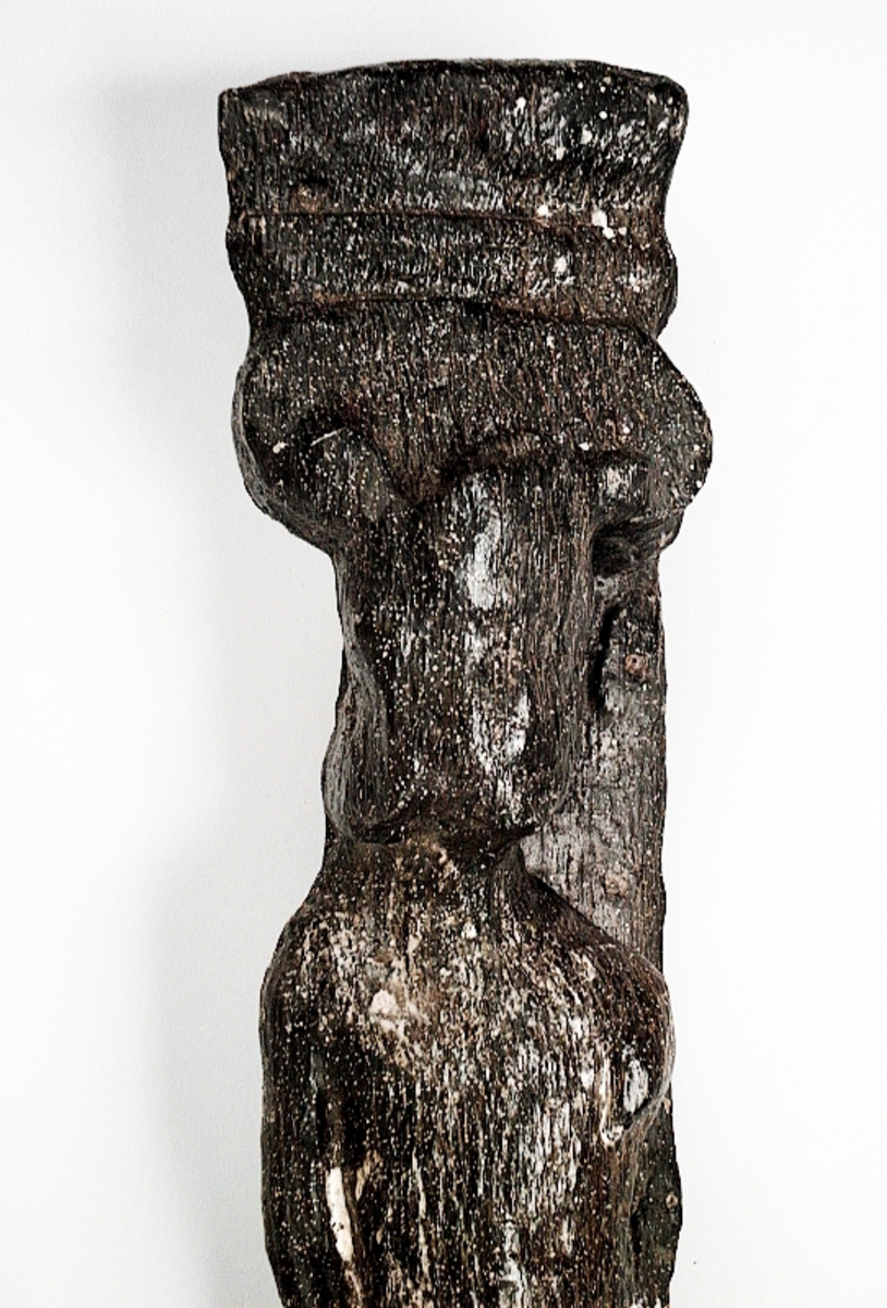 Skulpterad pilaster föreställande en herm eller möjligtvis en karyatid, under jonisk kapitäl.
Huvudet är vridet åt vänster samt prytt med en krona. Armarna hålls sträckta utefter sidorna.
Kroppen övergår i det med blad- och blomornamentik rikt sirade pilasterskaftet som smalnar av nedåt. Skaftet avgränsas genom en enkel vulst eller list från den rikt sirade och mjukt utbuktande piedestalen. Nedtill är piedestalen försedd med en bred bas. Skulpturens baksidan är slät.
Skulpturen är sliten.

Text in English: Sculptered pilaster with a Herm, or possibly a Caryatid, beneath an Ionic capital.
The head, with a crown, is turned to the left. The arms are streched out along the sides.
The body extends into the shaft of the pilaster, which tapers downwards and is richly decorated with leaf and flower ornamentation. A simple torus or batten separates the shaft from the richly decorated, softly bulging pedestal that has a broad base. The back of the sculpture is smooth.
The sculpture is worn.