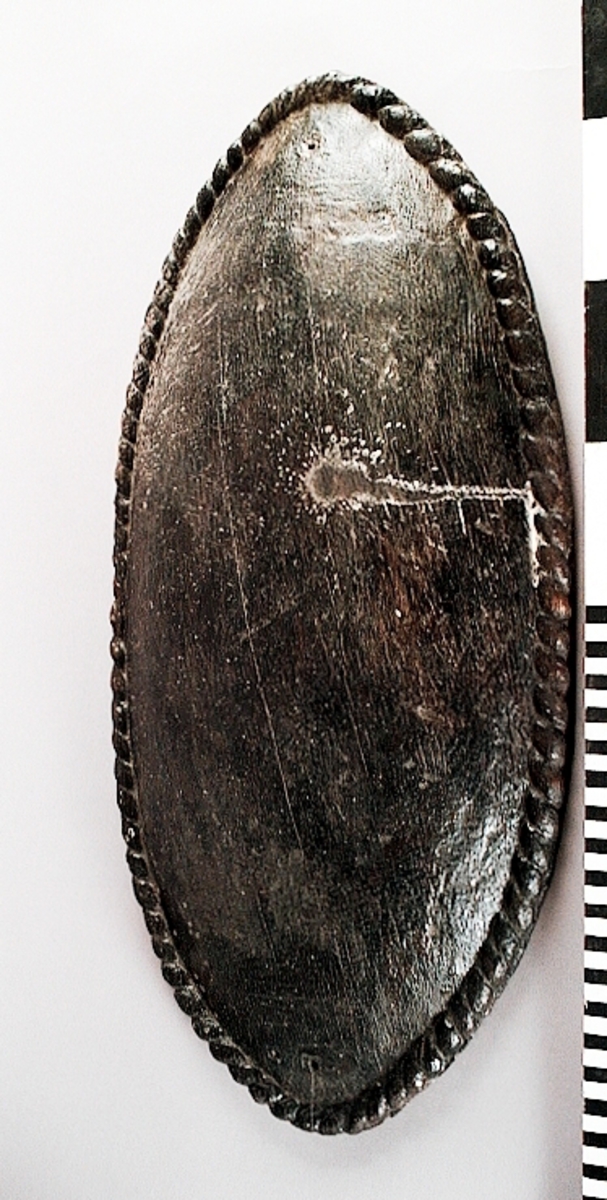 Skulpterad stor, oval och bukig sköld. Skölden pryds av en enkel och tunn kantbård, i form av ett snott band. Framsidan är slät, liksom den urgröpta baksidan. I den ena änden finns ett hål med ett runt märke efter spikhuvud. I den andra änden finns två hål, med fyrkantiga spår av spikhuvuden. Samtliga hål är placerade innanför kantbården.
Skölden är välbevarad, förutom några mindre sprickor.

Text in English: A sculpted large bulging oval shield.
The shield is decorated with a single cord edging, in the form of a slanting band. Front and back are smooth. The back is hollowed out.  At one end of the shield is a hole with the mark from a nail head, at the other end are two holes, with marks of square nail heads. All the holes are situated within the border.
The shield is well preserved, except for a few small cracks.
