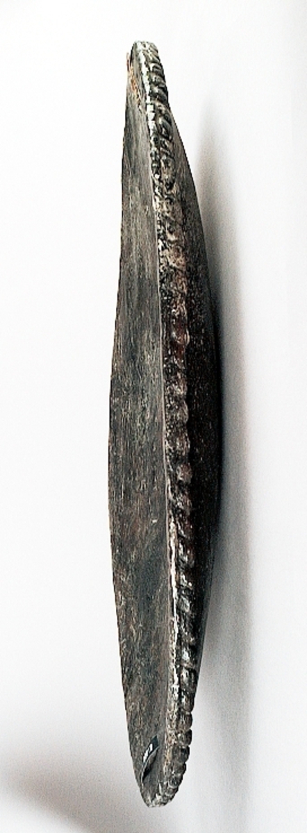 Skulpterad stor, oval och bukig sköld. Skölden pryds av en enkel och tunn kantbård, i form av ett snott band. Framsidan är slät, liksom den urgröpta baksidan. I den ena änden finns ett hål med ett runt märke efter spikhuvud. I den andra änden finns två hål, med fyrkantiga spår av spikhuvuden. Samtliga hål är placerade innanför kantbården.
Skölden är välbevarad, förutom några mindre sprickor.

Text in English: A sculpted large bulging oval shield.
The shield is decorated with a single cord edging, in the form of a slanting band. Front and back are smooth. The back is hollowed out.  At one end of the shield is a hole with the mark from a nail head, at the other end are two holes, with marks of square nail heads. All the holes are situated within the border.
The shield is well preserved, except for a few small cracks.