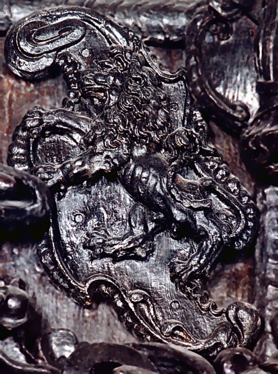 Skulpterad sköld, med upprätt, mot höger gående lejon. Lejonet är snidat i hög relief, med en heraldiskt stiliserad hållning. Sköldens kontur är rokokomässig, med kanter av varierande bredd och rik pärlbroskdekoration. Den vänstra kanten är något insvängd på mitten. Upptill avslutas skölden med en ellipsformad broskvolut. Baksidan är slät.

Text in English: Shield with upright, rightward-facing lion. The lion is carved in high relief and reproduced in perspective. It is seen in profile, and its posture is heraldically stylised. The shield has an almost rococo-like outline, with edges of varying width and rich earshell work decorations. The left-hand edge of the shield is somewhat narrowed at the middle.The top the shield is finished with an ellipse-shaped pearlwork scroll. The reverse side is smooth.