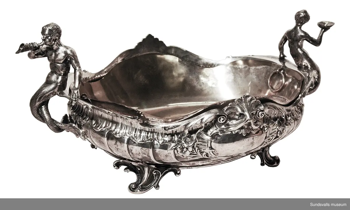 Två silverskålar på fötter med förgyllda löstagbara innerskålar, vilka är försedda med ringar att lyfta i. Skålarna är dekorerade med mytologiska figurer (sagofigurer?) och maskaroner. Gravyrfält. Nyrokoko
 SuM 2333:1 är 49 cm lång och 20 cm bred. G SuM 2333:2 är 48 cm lång och 21 cm bred. Skålarna är stämplade med 'C.G. HALLBERG', 'kattfoten' och Stockholms stads stämpel.