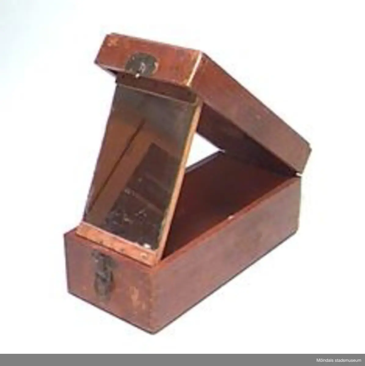 Låda i brunt trä. Öppnas på längden. Utfällbar spegel i locket. Material: även mässing.Nött och fläckig. Givare även: Wågdahl, Ann-Kristin, Sagbrov. 37, Lindome.