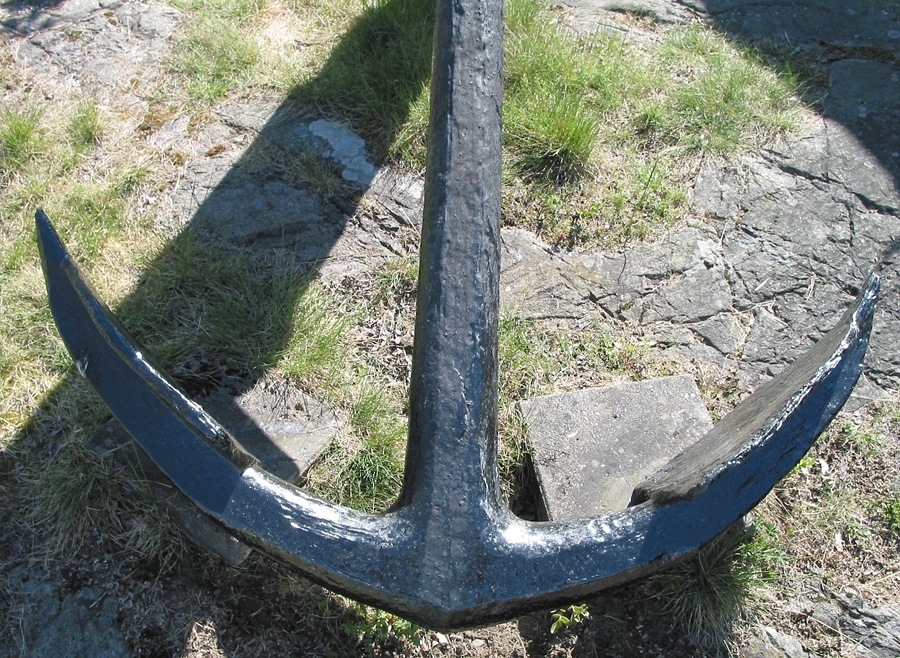 Stockankare. Trästock med fyra järnband samt en skyddskåpa i metall över ändträet på övre delen. Stockens två delar hålls ihop med fyra kilade trädymlingar. Stocken är på vissa partier mycket sliten. Schackel i röringen.