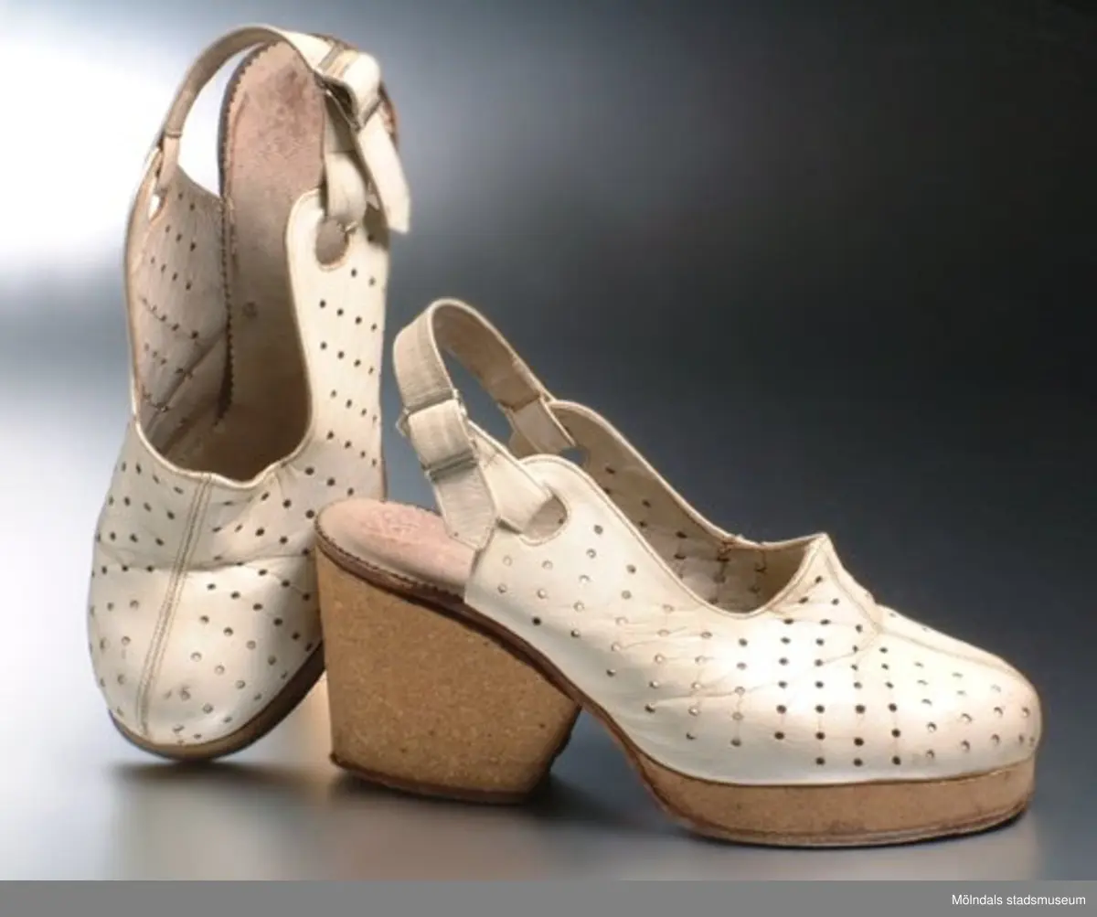 Vit sandalett med hög klack. Vid hälen sitter ett band med spänne. Har små "prydnadshål" ovanpå skon. Sandalen är gjord av skinn. Men sulan och klacken är gjord av trä.