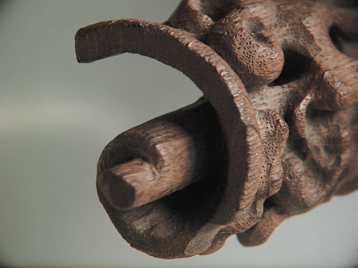Rullverksornament.
Skulpterat litet, spänneliknande ornament, dekorerat i broskverksstil.
Det långsmala ornamentet är format som ett stycke kraftigt upprullad bit näver eller läder, med konkav bak- eller undersida. På baksidan är ornamentet profilerat för en list. Broskdekoren på ovansidan är formad som en maskaron.
Ornamentet är mycket välbevarat.

Text in English: A small, buckle-like ornament, decorated in a style similar to earshell work.
The long narrow ornament is formed as a piece of tightly rolled bark or leather, with a concave back or underside so as to match a batten or the like. The reverse side is profiled to fit a moulding. The earshell work on the upper side is shaped as a mask.
The ornament is well preserved.