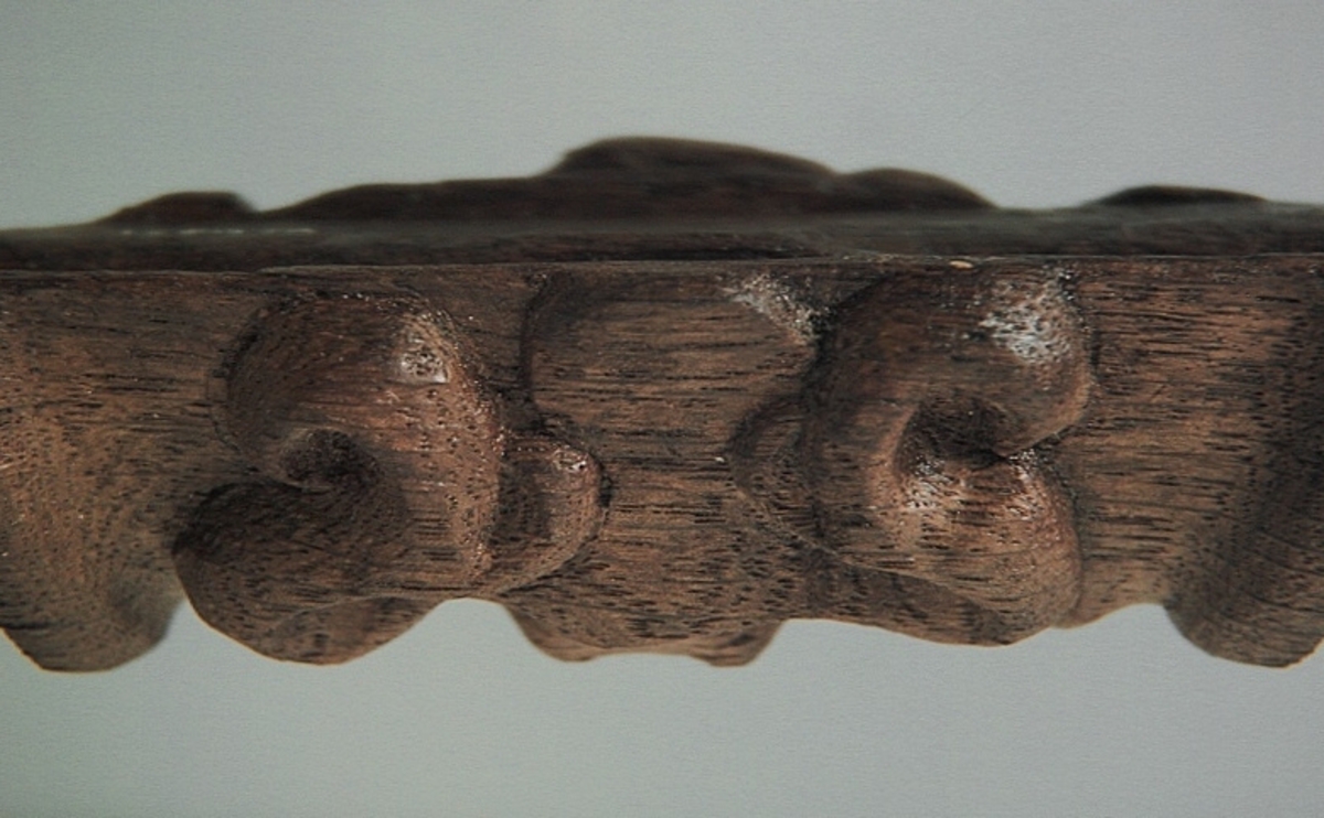 Rullverksornament.
Skulpterat litet, spänneliknande ornament, dekorerat i broskverksstil.
Det långsmala ornamentet är format som ett stycke kraftigt upprullad bit näver eller läder, med konkav bak- eller undersida. På baksidan är ornamentet profilerat för en list. Broskdekoren på ovansidan är formad som en maskaron.
Ornamentet är mycket välbevarat.

Text in English: A small, buckle-like ornament, decorated in a style similar to earshell work.
The long narrow ornament is formed as a piece of tightly rolled bark or leather, with a concave back or underside so as to match a batten or the like. The reverse side is profiled to fit a moulding. The earshell work on the upper side is shaped as a mask.
The ornament is well preserved.