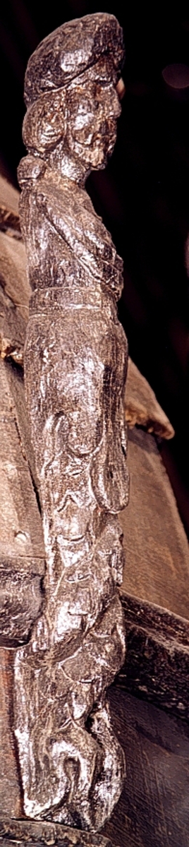 Skulpterad triton med turbanliknande lagerkrans på huvudet.

Huvudet har långt, ringlande hår samt stora och nedhängande mustascher. Snett över bröstet är en klädnad draperad. Klädnaden är fästad i en knut på höger axel. Kring livet sitter ett band med en oval.



Kroppens övergång till de dubbla, snodda fiskstjärtarna döljs av ett stort rundflikigt akantusblad. Huvudet är till största delen framställd som rundskulptur, dock något avplanad. Från nackhöjd och halvvägs ned på partiet med fiskstjärtarna är baksidan avplanad och slät - delvis något skålad - varefter det sitter ett snett urtag. Fotändan är vinkelformigt skålad vilket ger den en V-format tvärsektion, nedsänkningen är dock inte symmetriskt placerad. Nedtill böjer fotänden av inåt.

Skulpturen är mycket välbevarad.



Text in English: Sculpted Triton with a turban-like laurel wreath on its head.

The figure has a large pendulous moustache and long curly hair. There is a garment draped diagonally across the chest and fastened in a knot on the right shoulder. There is a girdle or belt round the waist with an oval centrepiece.



The transition between the body and the double fish tails is concealed by a large rounded acanthus leaf. The majority of the head is carved as a three-dimensional sculpture. The rear side surface has been planed smoothly.  The foot end has been hollowed out at an angle, giving it a V-shaped cross section, the hollowed-out section is not, however, symmetrically placed. The foot end curves inwards.

The sculpture is very well preserved.