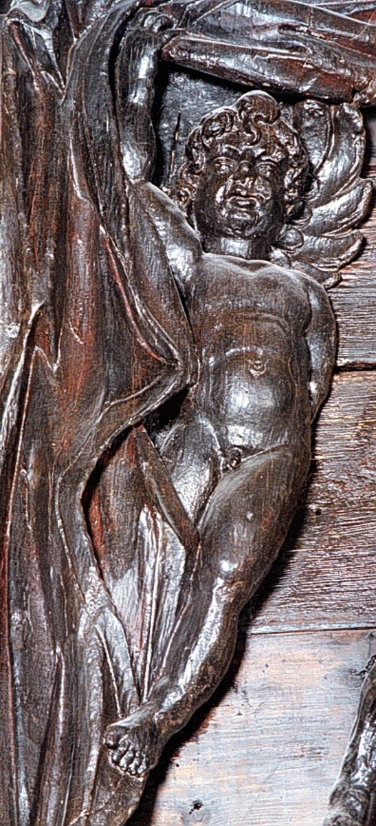 Putto samt större delen av ett draperi. Draperiet är kilformat med rak ytterkontur, och hålls undan av den svävande putton. Skulpturens baksida är slät.

Text in English: Putto and the larger part of a drapery. The drapery is wedge-shaped with straight outer contour, and is held back by the hovering putto. The reverse side is smooth.