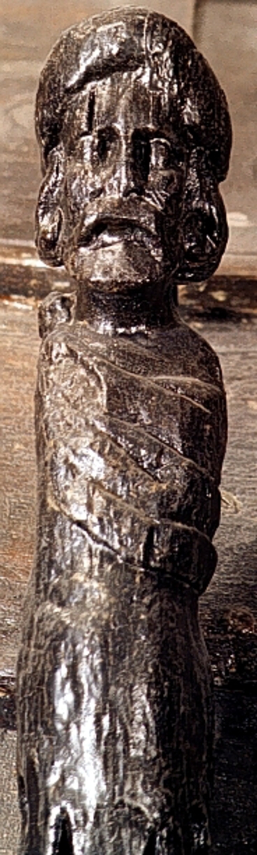 Skulpterad Triton med tjock lagerkrans på huvudet.
Figuren har stora yviga mustascher och långt, lockigt hår. Snett över bröstet är en klädnad draperad. Klädnaden är hopfäst på vänster axel.

Övergången till de snodda fiskstjärtarna döljs av ett stort, flikigt blad vars mittflik är förtjockad och upprullad. Skulpturens fotända är tapp- eller kilformad och tillplattad från sidorna. Tritonens huvud är framställt som rundskulptur. Från axlarna och nedåt är baksidan slät och plan. I höjd med övergången till stjärtpartiet finns ett kraftigt urtag. Nedtill bryter baksidans fotända av inåt.

Text in English: Sculpted Triton with thick laurel wreath on its head.
The figure has a large bushy moustache and long curly hair. A garment is draped across the chest and fastened on the left shoulder.

The transition between the upper body and fish tails is concealed by a large leaf, the middle segment of which is thickened and rolled up. The foot end of the sculpture has a flat end, seen from the side, and is wedge-shaped. The Triton''s head is carved as a three-dimensional sculpture. The rear side is smooth. There is a deep groove on the rear side at the transistion point between the male torso and the fish tails.